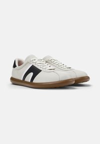 Un paio di sneaker bianche basse con accenti neri sui lati, lacci bianchi e suole in gomma marrone su uno sfondo chiaro e semplice.