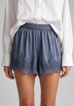Kvinde iført løstsiddende blå satinshorts med blondekant og en hvid langærmet knap-op-shirt stukket ind i taljen.