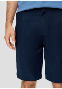 Shorts bleu marine en tissu texturé, avec un devant plat et des poches latérales, arrivant juste au-dessus du genou. Main positionnée à proximité.