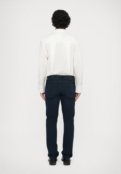 Λευκό πουκάμισο με μακριά μανίκια και σκούρα μπλε τζιν, φαίνεται από πίσω. Το πουκάμισο έχει μια διακριτική γυαλάδα, ενώ τα τζιν έχουν κλασική εφαρμογή και ορατές τσέπες.