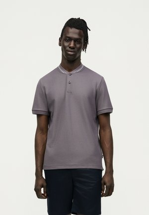 Νεαρός άνδρας με σκούρα πλεξούδες, ντυμένος με γκρι κοντομάνικο πόλο και μαύρα σορτς, στέκεται μπροστά σε απλό ανοιχτόχρωμο φόντο.