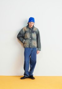 Denimjakke med falmede grønne og beige nuancer, to brystlommer, parret med blå jeans og sorte sko, toppet med en blå hue.