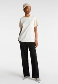 Short-sleeve t-shirt in cream and beige, made of cotton, featuring a round neckline and subtle logo detail. Paired with black wide-leg pants.