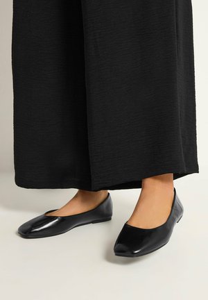 Zwarte leren ballerina's gedragen met brede zwarte gestructureerde broek op een lichte vloer achtergrond.