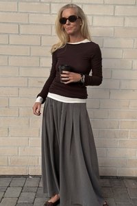 Langer grauer Faltenrock kombiniert mit einem enganliegenden dunkelblauen Oberteil mit weißem Kragen, schwarzen Sonnenbrillen und einer Kaffeetasse haltend.