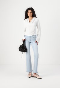 Haut à manches longues blanc avec col drapé, jeans évasés bleu clair, chaussures en cuir beige et un sac à cordon noir à motif floral.