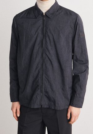 Mand iført en mørk koksgrå lynlåstrøje over en beige rullekrave og sorte bukser, stående mod en ensfarvet baggrund.