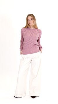 Vrouw met lang blond haar, gekleed in een mauve textuur trui en wijd uitlopende witte broek, staand met handen in de zakken.