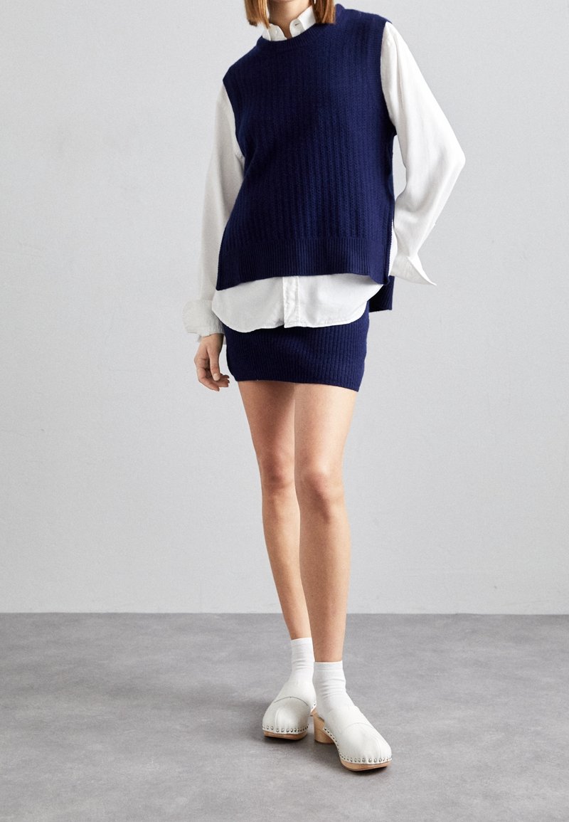 Chaleco de punto acanalado azul marino superpuesto sobre una camisa blanca con cuello, combinado con una falda corta azul marino a juego y zuecos blancos con suelas de madera.