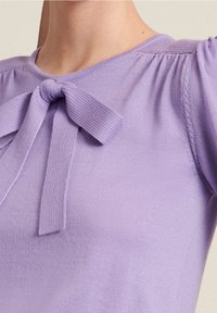 Lila gebreide top met een ronde hals, voorzien van een strikdetail middenvoor en getextureerde accenten op de schouders. Zachte, lichte stof.