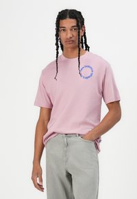 Pink cotton t-shirt with short sleeves, featuring a blue circular logo with text. Paired with light gray jeans, the look is casual and sporty.