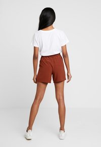 Pantaloni corti marrone con vita elasticizzata abbinati a una maglietta bianca, visti da dietro. Scarpe da ginnastica bianche completano il look informale. Tessuto morbido.