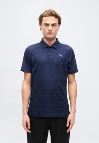 Jeune homme debout face à l'avant, portant un polo Puma bleu marine à motifs et un pantalon noir, sur un fond blanc uni.