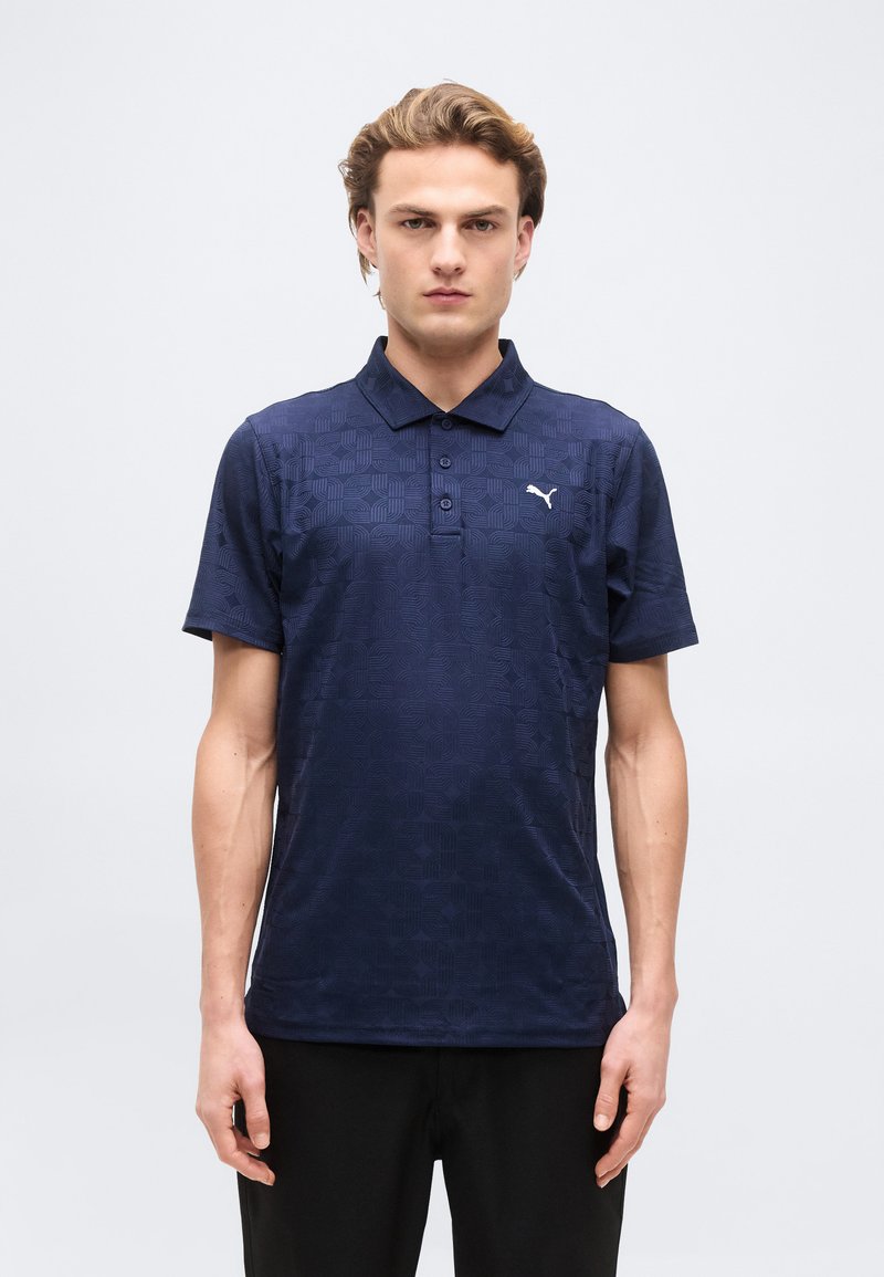 Jeune homme debout face à l'avant, portant un polo Puma bleu marine à motifs et un pantalon noir, sur un fond blanc uni.