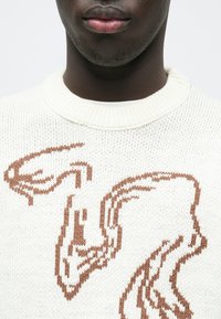 Kremasto bež pulover iz pletenine z teksturiranim vzorcem v topli rjavi barvi, na sprednji strani pa izstopajo abstraktne oblike.