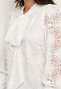 Vit spetsblus med intrikata blommönster, en snörning vid halsen och långa, utsvängda ärmar med en genomskinlig textur.