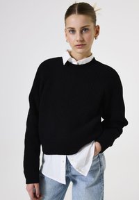 Jongeman met haar opgestoken, draagt gouden creolen, een zwarte gebreide trui over een witte blouse en lichtblauwe spijkerbroek met haar hand in haar zak.