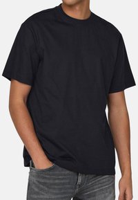 Schwarzes Baumwoll-T-Shirt mit kurzen Ärmeln, rundem Halsausschnitt und lockerer Passform. Geschmeidige Textur; getragen mit grauen Jeans.