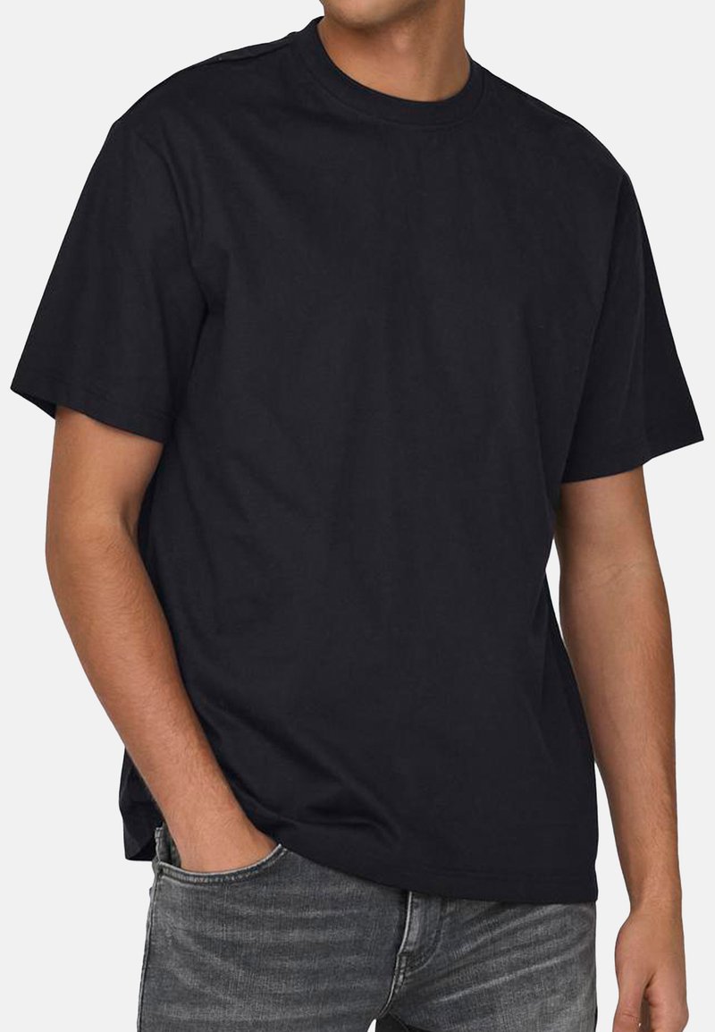 Schwarzes Baumwoll-T-Shirt mit kurzen Ärmeln, rundem Halsausschnitt und lockerer Passform. Geschmeidige Textur; getragen mit grauen Jeans.