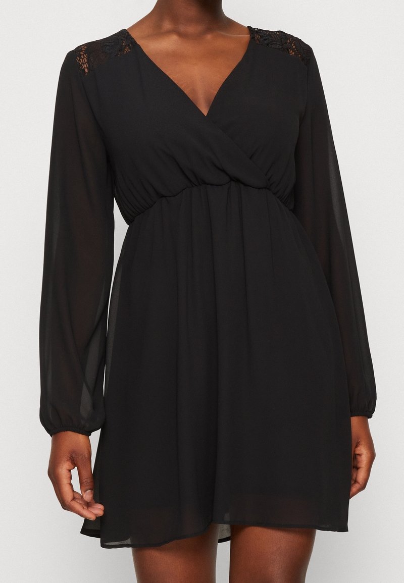 Robe noire avec manches longues transparentes, encolure en V, taille froncée et détails en dentelle aux épaules, portée par une personne debout.