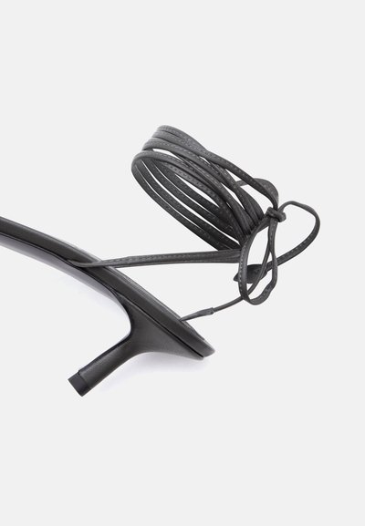 Μαύρο σανδάλι με λεπτούς λουράκια πολλαπλών βρόχων στον αστράγαλο και κοντό, λεπτό τακούνι σε λευκό φόντο.