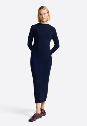 Woman with short blonde hair wearing a fitted navy ribbed long-sleeve midi dress and burgundy lace-up shoes, standing with hands behind back.