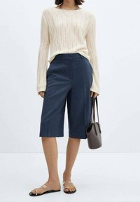 Vaalean kermanvärinen ribbineulottu pitkähihaisen villapaita, yhdistetty keskipitkiin tummansinisiin culottes-housuihin. Yhdistetty läpinäkyviin sandaaleihin ja pieneen tummanväriseen käsilaukkuun.