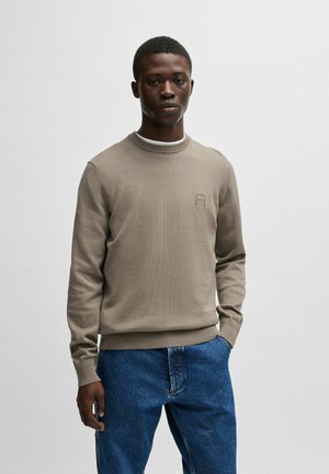 Maglione beige con collo rotondo e polsini a coste, dotato di una piccola patch con logo sul petto. Indossato con jeans in denim blu.