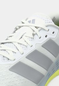 Άσπρο αθλητικό παπούτσι με διαπνεόμενο επάνω μέρος από δίχτυ, γκρι λογότυπο με τρεις ρίγες, υφασμένα πάνελ και λεπτομέρειες στο πέλμα σε λάιμ πράσινο χρώμα.