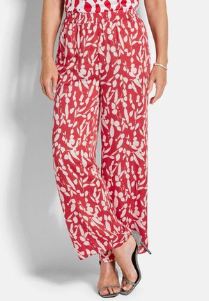 Femme portant un pantalon à jambes larges rouge et blanc à motifs et des sandales argentées à talons hauts, debout devant un fond uni.