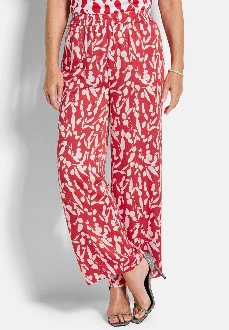 Femme portant un pantalon à jambes larges rouge et blanc à motifs et des sandales argentées à talons hauts, debout devant un fond uni.