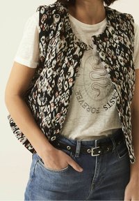 Gestepptes Weste mit einem schwarz-weiß-orangeblühenden Muster, getragen über einem hellen Hemd, kombiniert mit Jeans und einem schwarzen Gürtel.