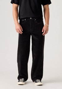 Homme portant un pantalon large en velours côtelé noir, un t-shirt noir, des baskets noires et blanches, debout devant un fond neutre.