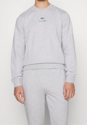 Sweatshirt gris avec col rond, poignets et ourlet côtelés, arborant un petit logo Lacoste vert sur la poitrine. Tissu en mélange de coton doux et lisse.