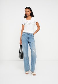 Eine Frau trägt ein weißes T-Shirt mit dem Guess-Logo, helle blaue, zerrissene Jeans, weiße Sandalen und hält eine schwarze Handtasche vor einem weißen Hintergrund.