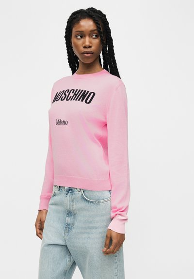 Femme aux longues tresses portant un pull rose Moschino Milano et un jean bleu clair, regardant légèrement vers sa gauche.