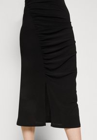 Czarna spódnica midi z marszczeniami po bokach i centralnym rozcięciem, noszona poniżej kolan na jednolitym jasnym tle.