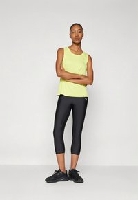 Light yellow sleeveless athletic top paired with black mid-length leggings; model wears black sneakers, arms crossed, neutral background.