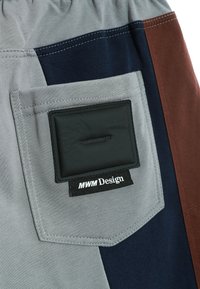 Tela gris con paneles en azul marino, gris y marrón. Presenta un gran bolsillo trasero con un parche rectangular negro etiquetado como "MWM Design."