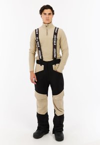 Skikleding met een beige lange mouwen top en twee-kleurige skibroek in zwart en beige met verstelbare bretels en meerdere zakken.