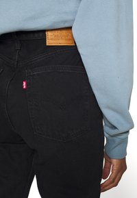 Jeans in denim nero con un'etichetta rossa Levi's ben visibile sulla tasca posteriore. La persona indossa un felpa azzurro chiaro, che copre parzialmente i jeans.