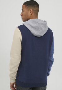 Sudadera azul marino con capucha gris claro, mangas beige y puños acanalados. Textura suave, diseño casual, con un bolsillo canguro.