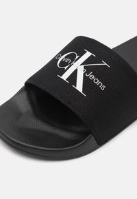 Svart slide sandal med en stoffstropp som har hvit "Calvin Klein Jeans" logo. Teksturt såle med en glatt overflate. Lett design.