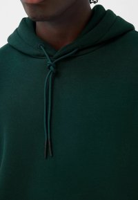 Felpa verde realizzata in morbido tessuto, con cappuccio regolabile dotato di dettaglio annodato e punte nere, texture liscia e vestibilità comoda.