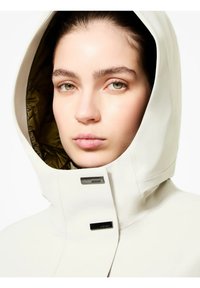 Giacca leggera di colore crema con cappuccio e fodera interna lucida verde oliva, dotata di accessori neri e design snello.