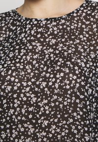 Haut en tissu noir transparent orné de motifs floraux blancs, avec un col rond et une finition légère et texturée.