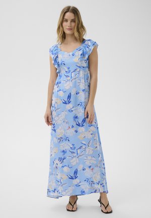 Nő kék, virágmintás maxi ruhában, fodros ujjú és négyzet alakú nyakkivágással, fekete, pántos szandállal, egyszínű háttér előtt állva.