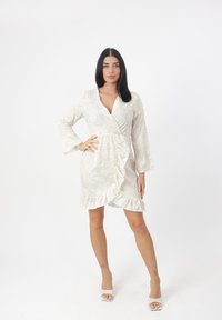 Floral Wickelkleid aus cremefarbenem Stoff mit langen Ärmeln, V-Ausschnitt, Rüschensaum und seitlichem Bindeband. Kombiniert mit weißen offenen High Heels.