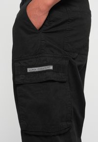 Zwarte cargobroek met een zijzak met een grijs label met de tekst "ALPHA INDUSTRIES." De stof heeft een soepele textuur en een gestructureerde pasvorm.