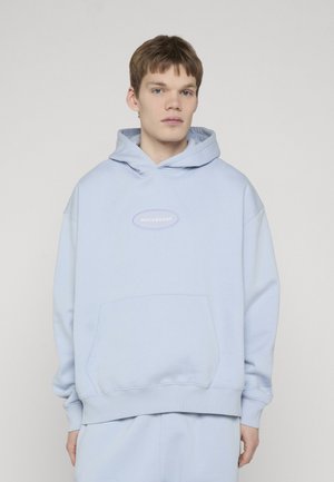 Joven con sudadera con capucha Rocawear azul claro con bolsillo delantero, de pie contra un fondo liso y claro.